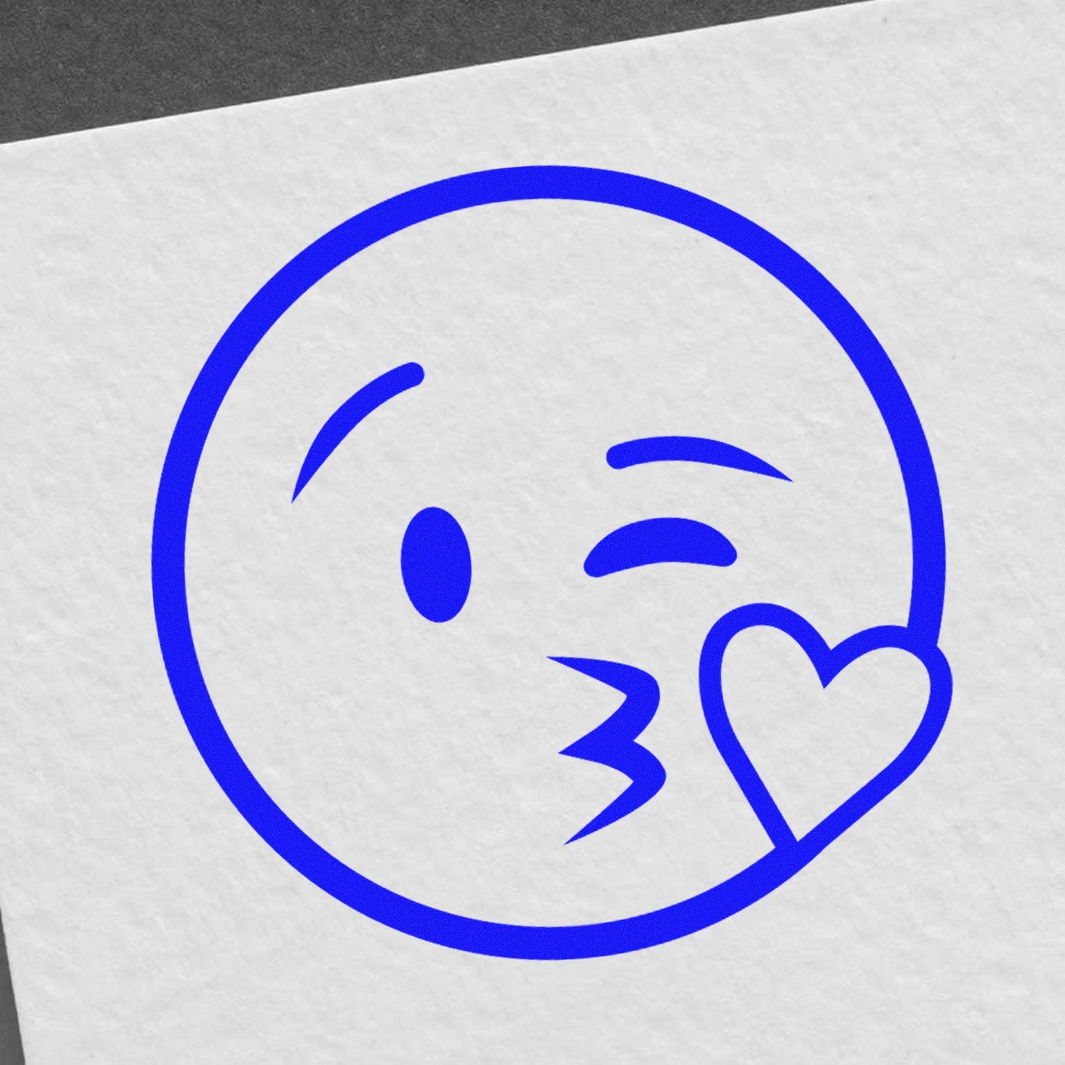 Self-Inking Blowing a Kiss Emoji Stamp in blue ink on white paper, featuring a winking face with a heart. Perfect for adding a playful touch to notes and crafts.