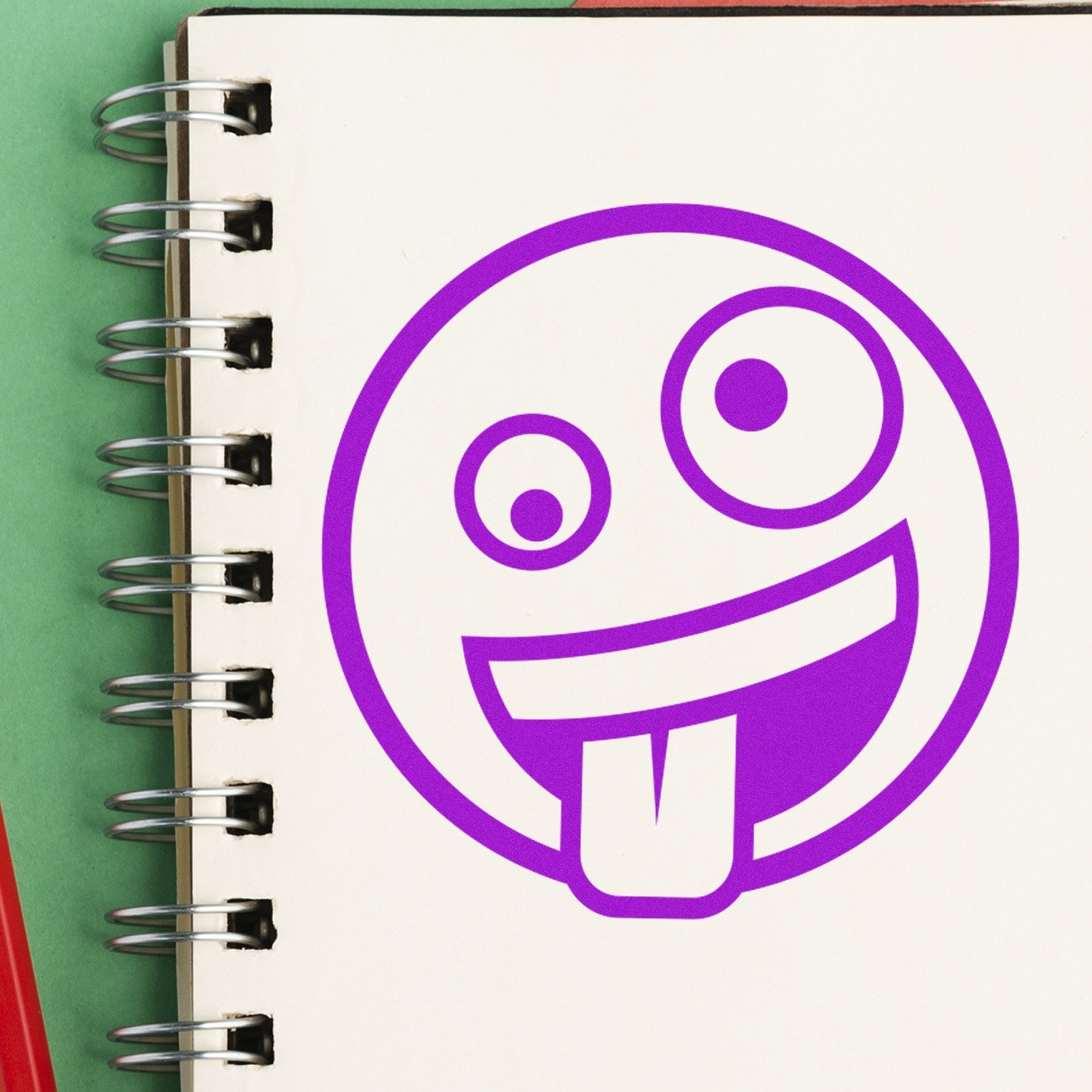 Wood Handle Zany Grin Emoji Rubber Stamp on a spiral notebook, featuring a playful purple emoji with a winking eye and tongue out. Perfect for adding fun to crafts and stationery.