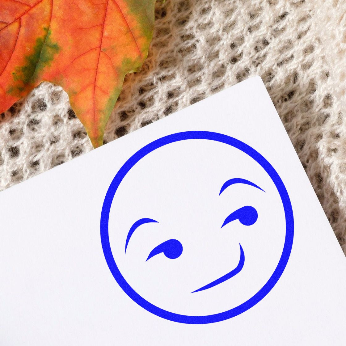 Wood Handle Smug Mug Reaction Rubber Stamp on textured fabric with autumn leaf, featuring a blue smug face design. Perfect for adding personality to crafts and stationery.