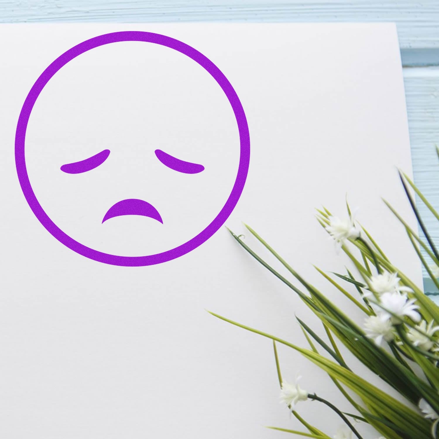 Wood Handle Sad Sammy Emojicons Rubber Stamp imprint on white paper, featuring a purple sad face emoji, next to green foliage on a light blue wooden surface.