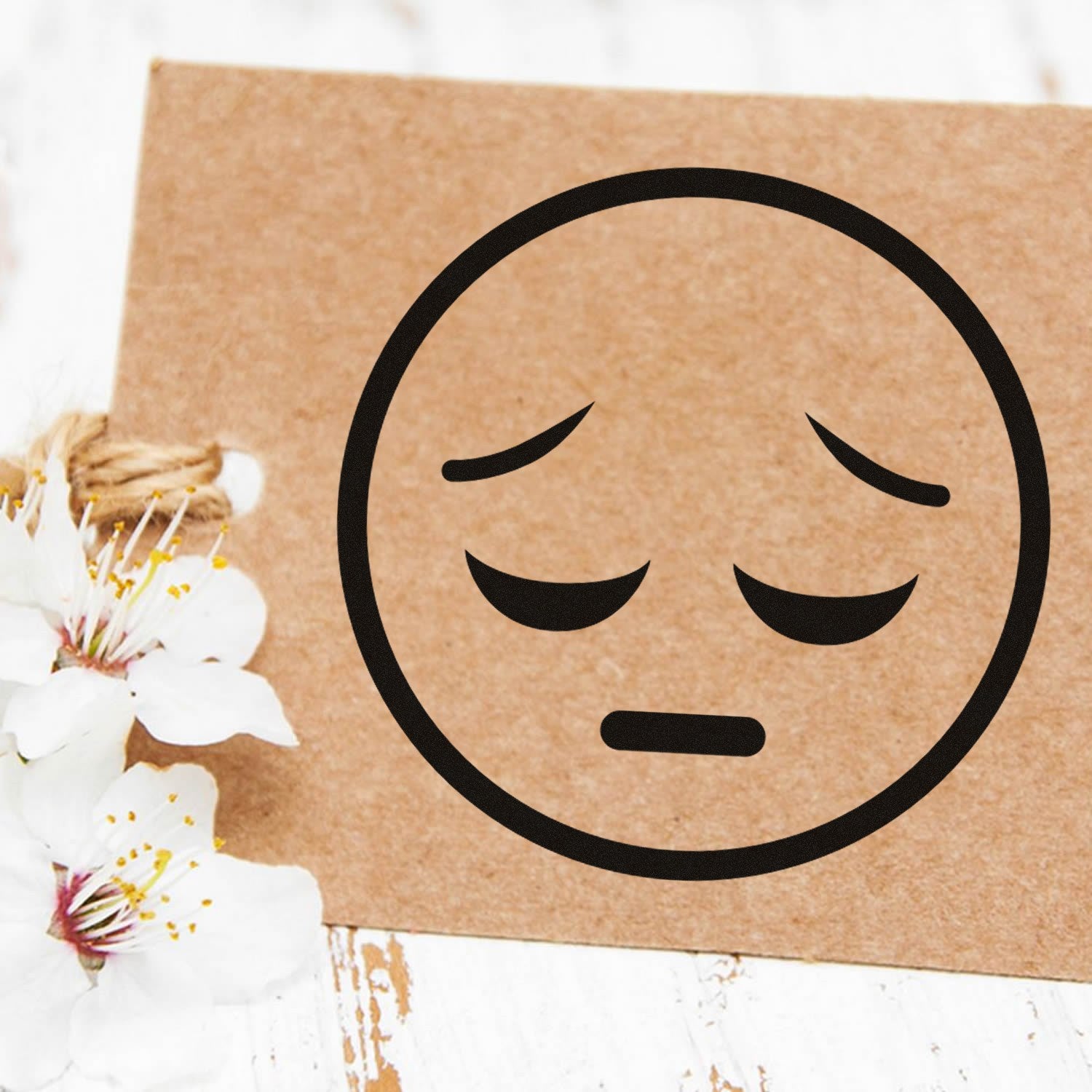 Self-Inking Silent Sorrow Emoji Stamp on brown card with white flowers, showcasing a sad face design. Perfect for adding a touch of emotion to your crafts and stationery.