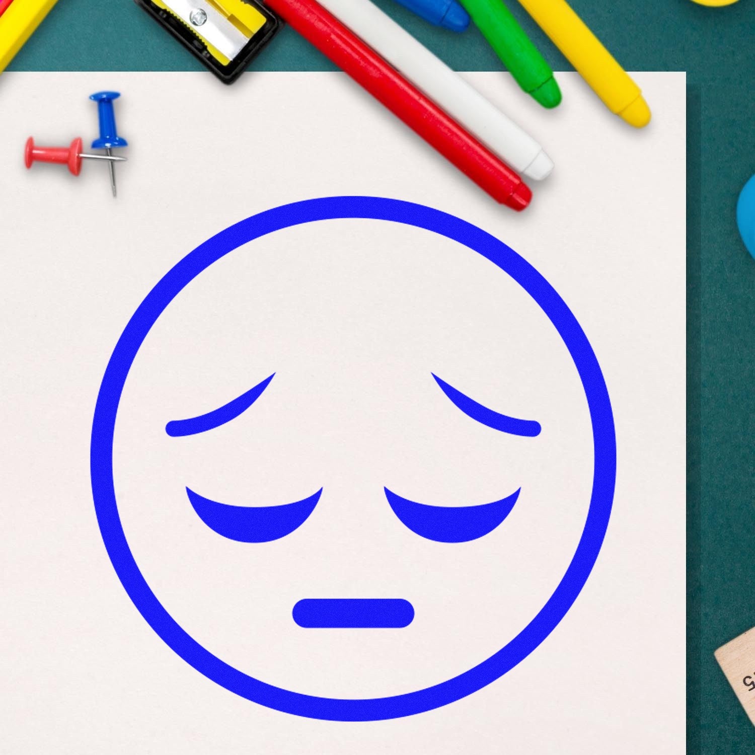 Self-Inking Silent Sorrow Emoji Stamp on paper, featuring a blue sad face. Surrounded by colorful pens and push pins, ideal for adding expressive touches to documents and crafts.