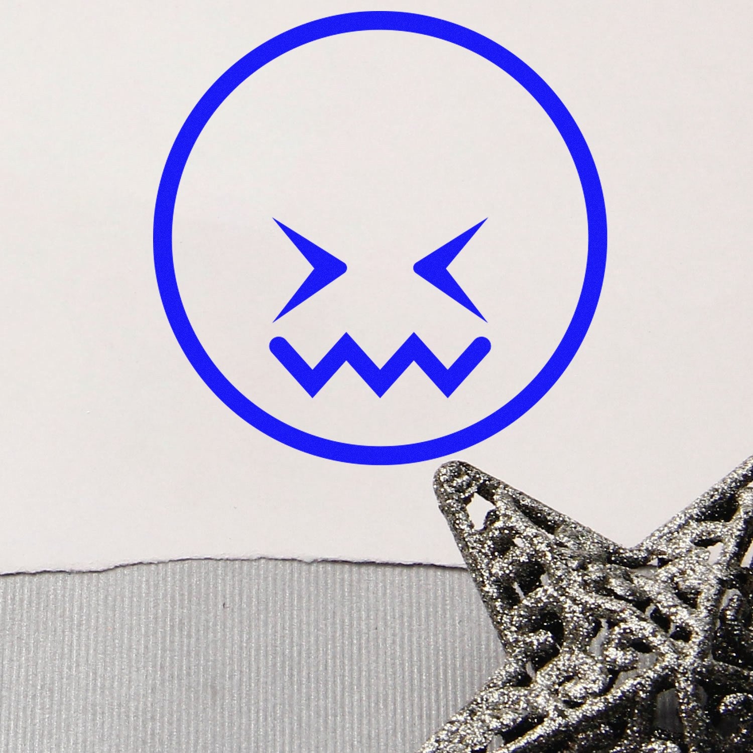 Self-Inking Zigzag Worry Emoji Stamp with a blue zigzag mouth and slanted eyes on white paper, next to a decorative silver star. Perfect for adding a playful touch to documents and crafts.