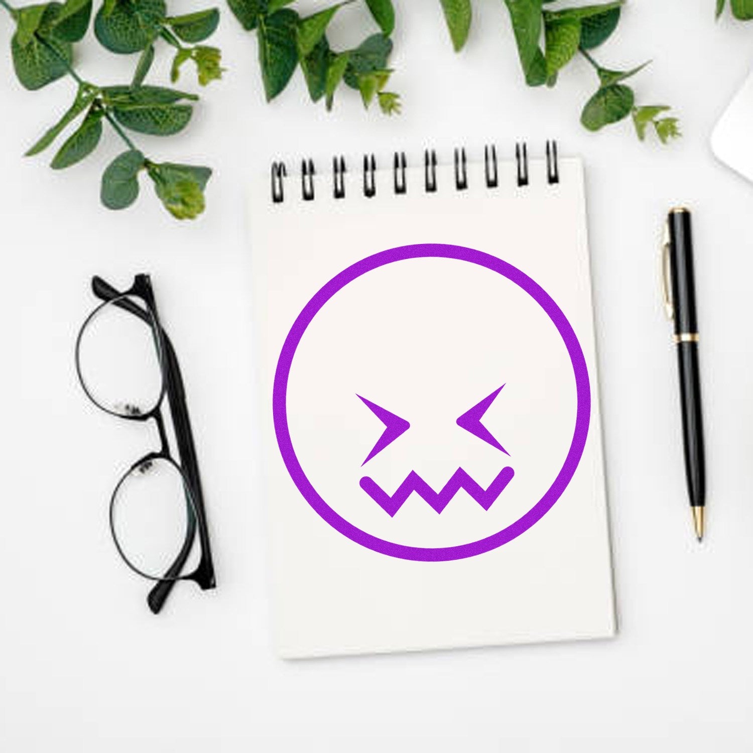 Self-Inking Zigzag Worry Emoji Stamp on notepad with purple zigzag mouth and eyes, surrounded by glasses, pen, and green leaves.