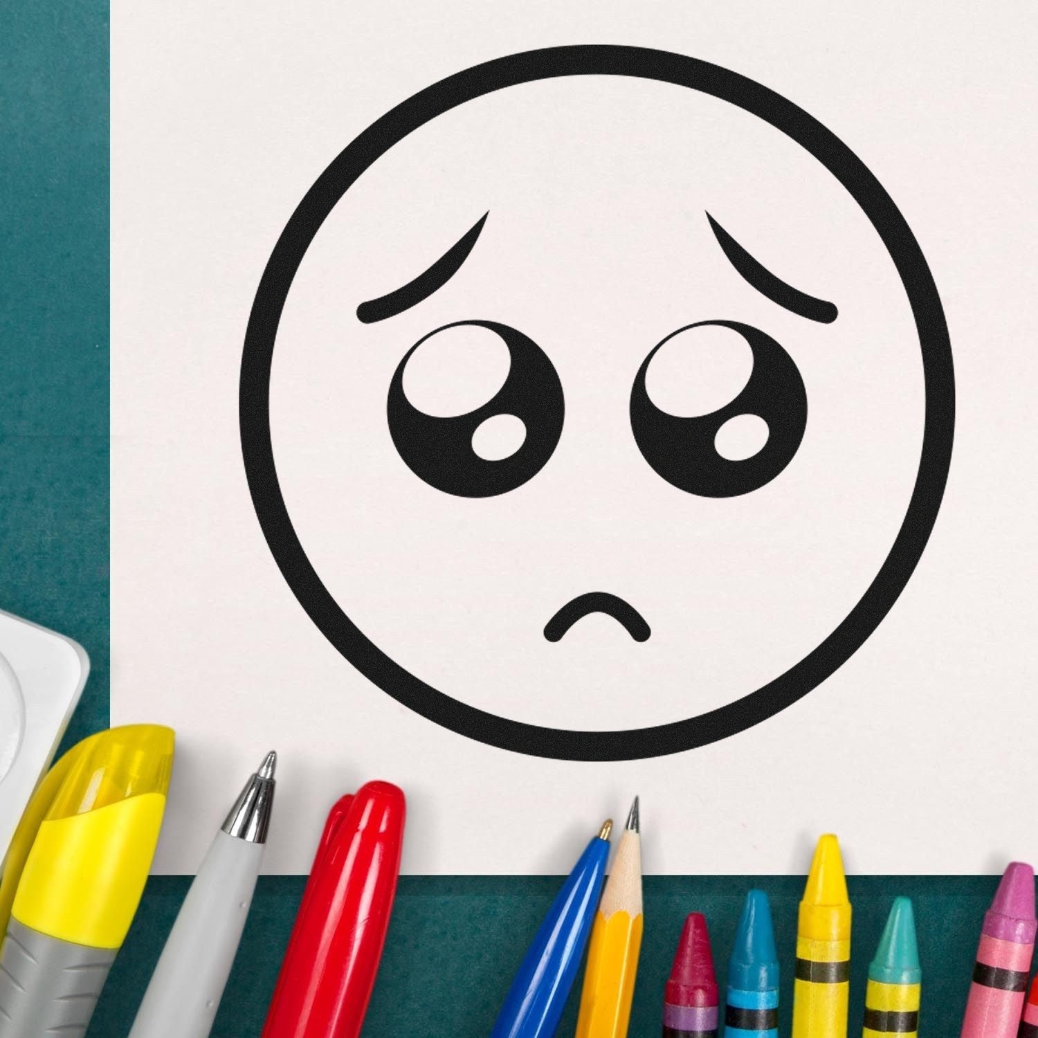 Self-Inking Puppy Eyes Emoji Stamp on paper with colorful pens and crayons, showcasing a cute, sad emoji design. Perfect for adding fun expressions to crafts and notes.