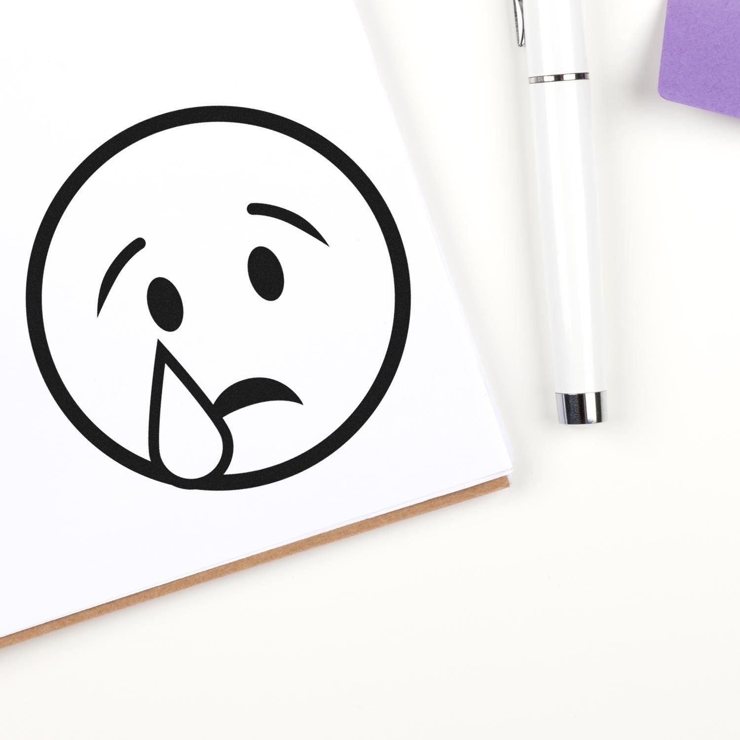 Self-Inking Teary Timmy Emoji Stamp on white paper, showing a crying face design. Perfect for adding a playful touch to notes and crafts. Pen and purple sticky note in the background.