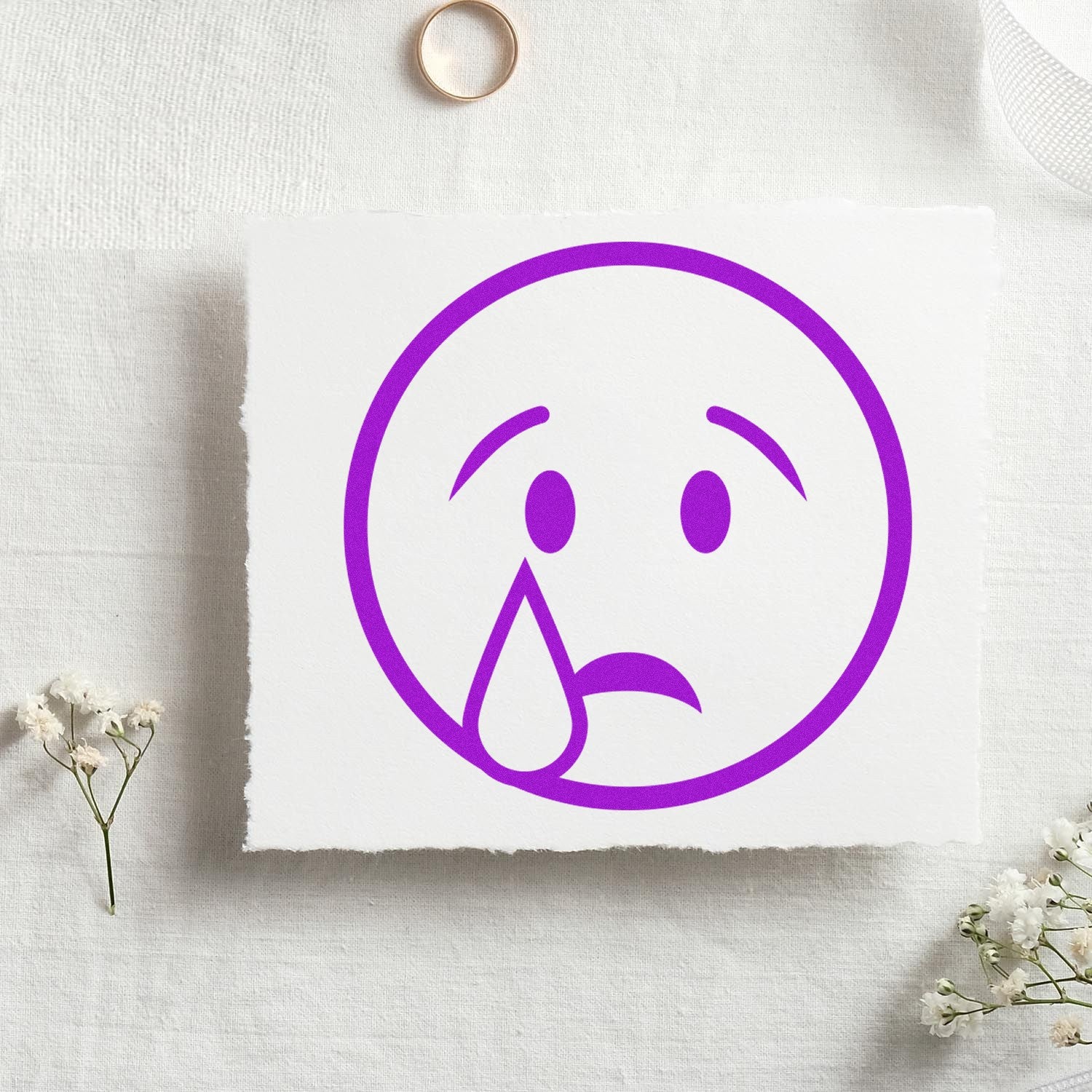 Wood Handle Teary Timmy Emoji Rubber Stamp featuring a purple teary-eyed emoji design on white paper, surrounded by delicate flowers and a gold ring on a textured surface.