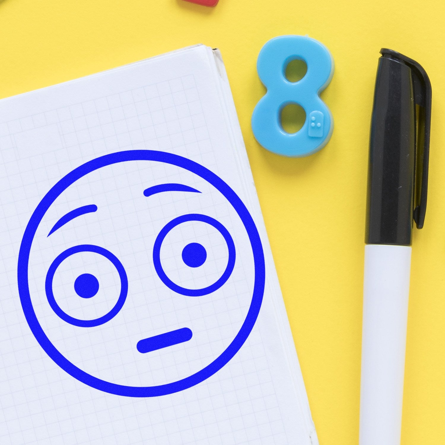 Wood Handle Wide-Eyed Wally Pictogram Rubber Stamp on yellow background, featuring a blue wide-eyed face imprint on white paper, next to a black pen and blue number eight.