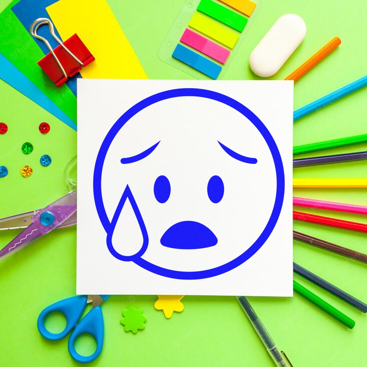 Self-Inking Tearful Timmy Facial Expressions Stamp on green background with colorful stationery, featuring a blue crying face design. Perfect for adding expressive details to crafts and projects.