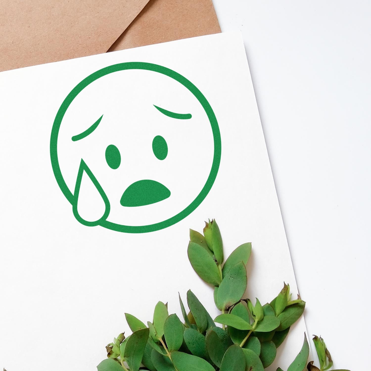 Self-Inking Tearful Timmy Facial Expressions Stamp creating a green crying face on white paper, surrounded by green leaves and a brown envelope.