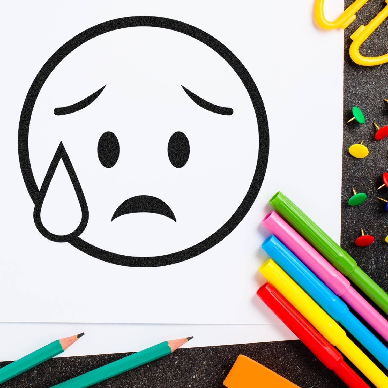 Self-Inking Weepy Wanda Emojicons Stamp on paper, showing a sad emoji with a tear. Surrounded by colorful pens and paperclips, perfect for adding expressive touches to crafts and documents.