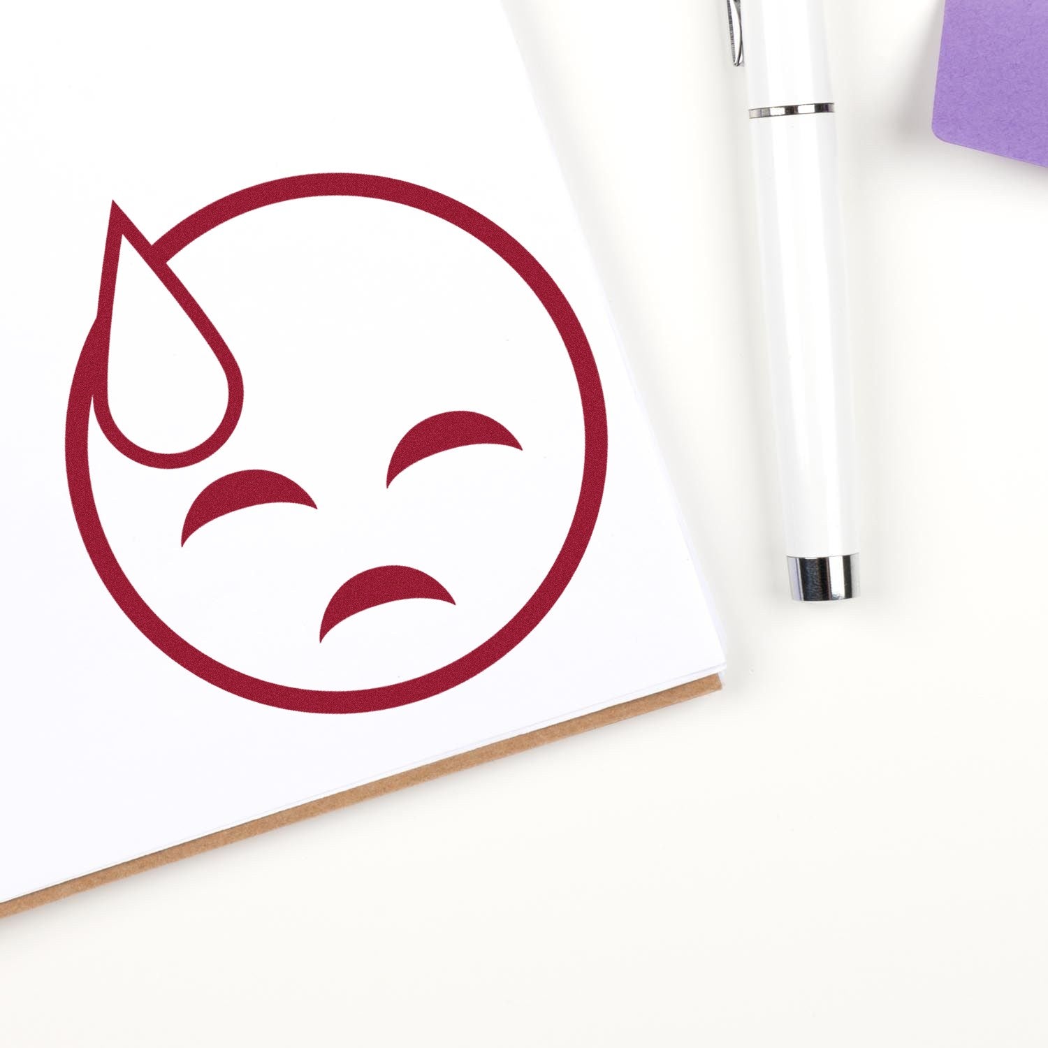 Self-Inking Sweaty Sadface Emoji Stamp imprint on paper, featuring a red sad face with a sweat drop. Includes a pen and purple notepad in the background.