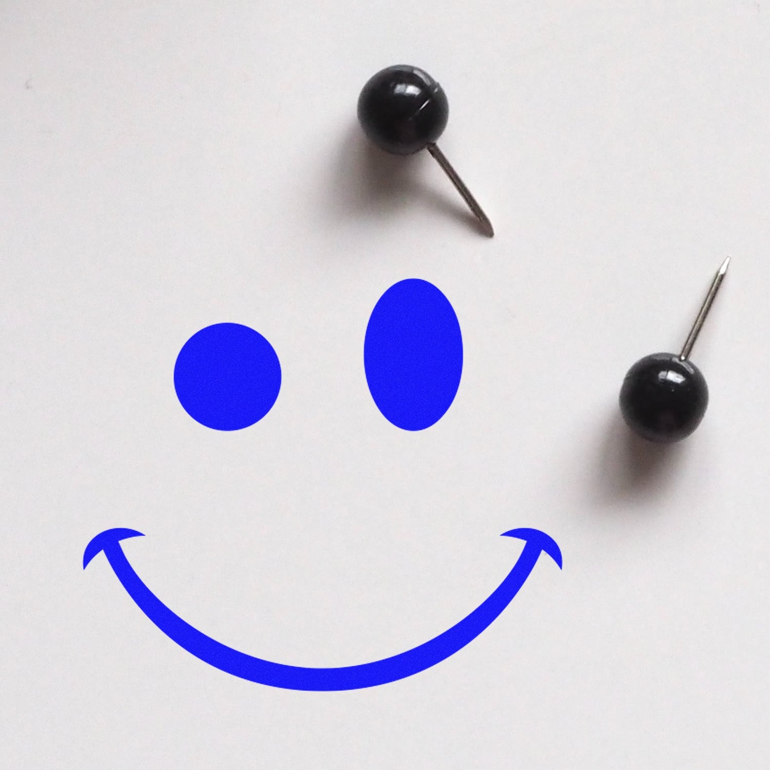 Self-Inking Mischievous Grin Emoji Stamp creating a playful blue smiley face on white paper, with two black push pins as eyes. Perfect for adding fun to documents and crafts.