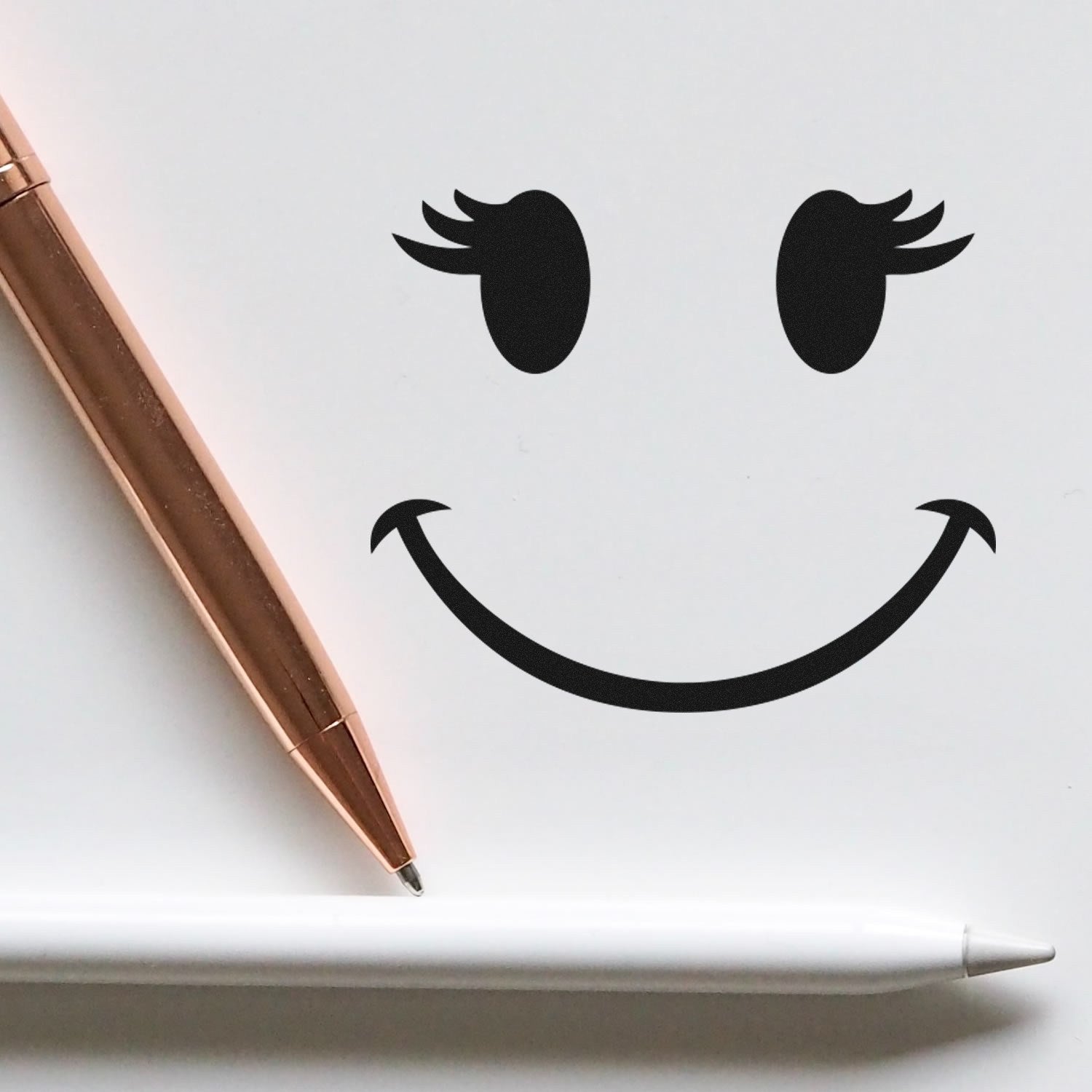 Self-Inking Fluttery Lashes Emoji Stamp on white paper, creating a smiling face with fluttery lashes. Includes a rose gold pen and a white stylus for scale. Perfect for adding fun to notes.