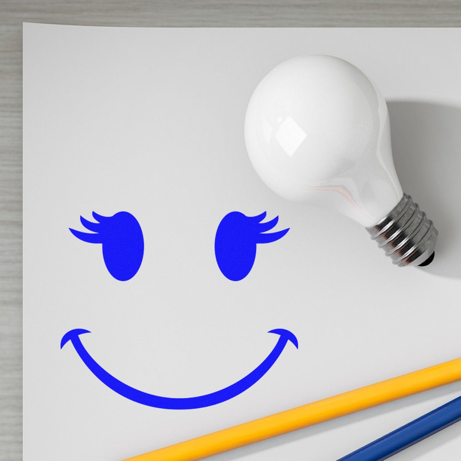 Self-Inking Fluttery Lashes Emoji Stamp on paper, creating a blue smiling face with lashes. Includes a light bulb and pencils for scale. Perfect for adding fun, expressive designs.