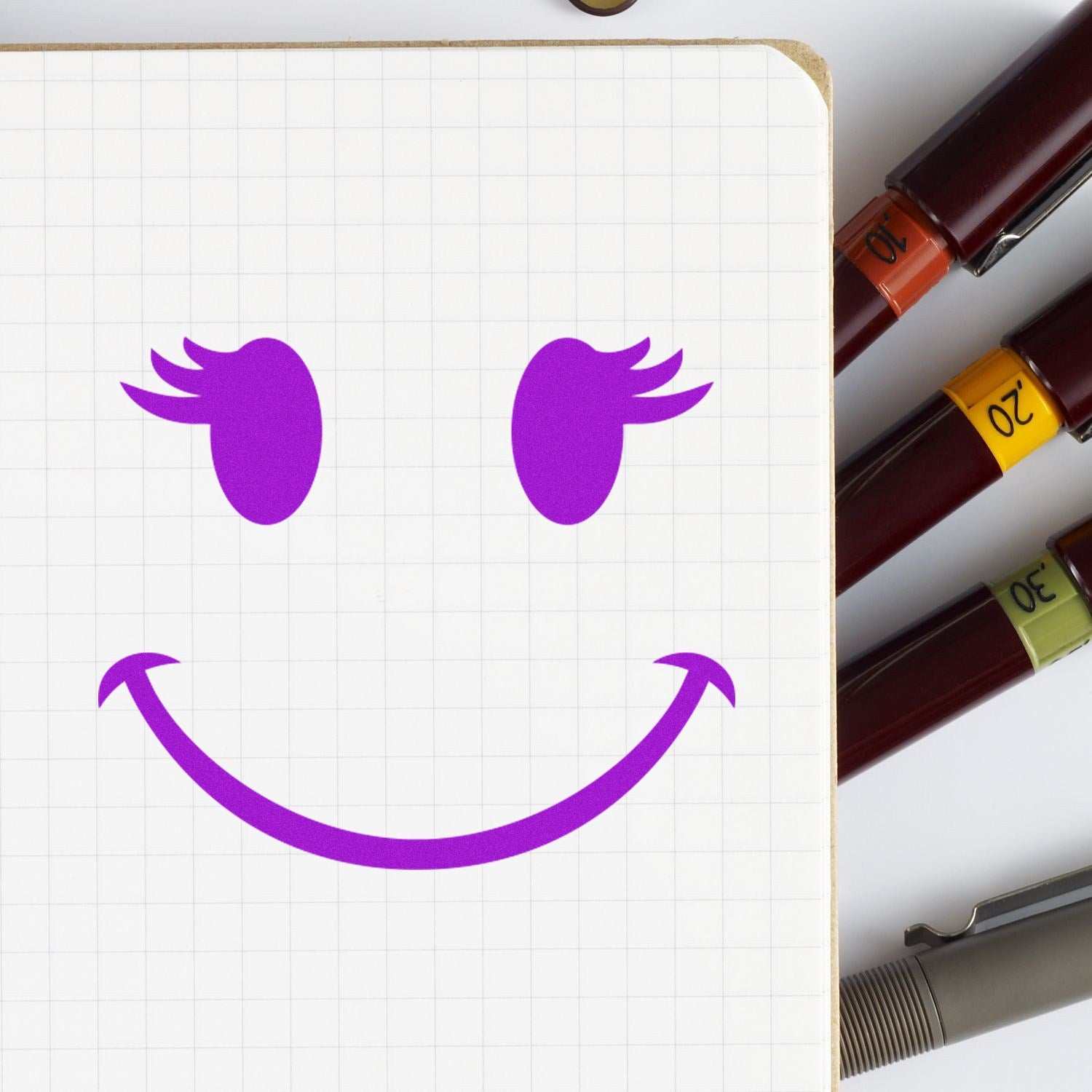 Wood Handle Fluttery Lashes Emoji Rubber Stamp on grid paper, creating a purple smiling face with fluttery lashes. Surrounded by colorful pens, showcasing its playful design and crafting use.