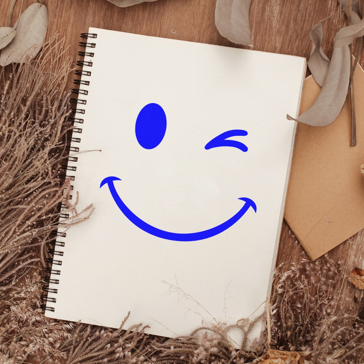 Wood Handle Winky Smiley Emoji Rubber Stamp imprint on a notebook, surrounded by dried leaves on a wooden surface.