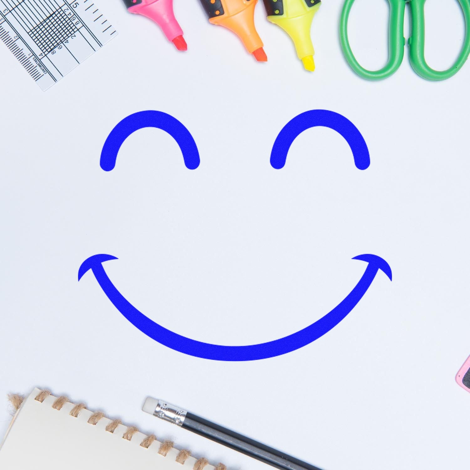 Wood Handle Giggly Smiley Emoji Rubber Stamp on white paper, surrounded by colorful highlighters, scissors, and a notebook, showcasing its cheerful blue smiley face imprint.