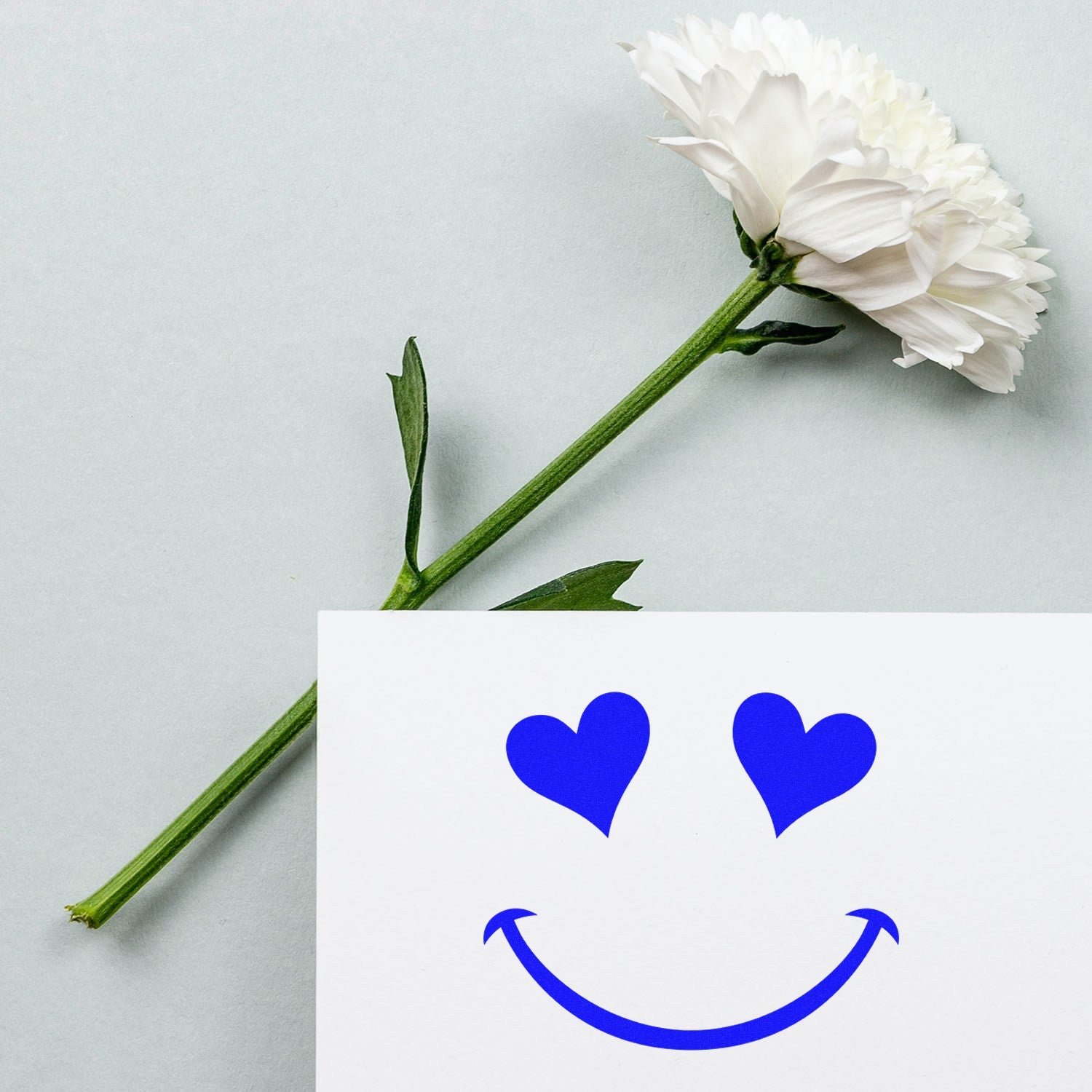 Self-Inking Heart Eyes Delight Emoticon Stamp in use, featuring a blue heart-eyed smiley face on paper, next to a white flower on a light gray background. Perfect for crafts and stationery.