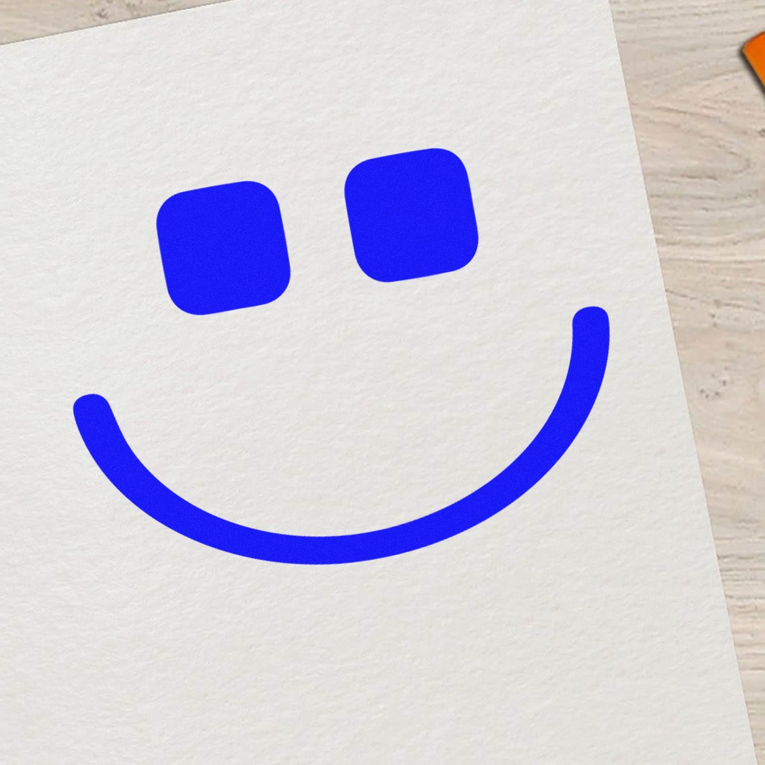 Wood Handle Geeky Grin Emotes Rubber Stamp on paper, featuring a bold blue smiley face with square eyes. Perfect for adding a playful touch to crafts and stationery.