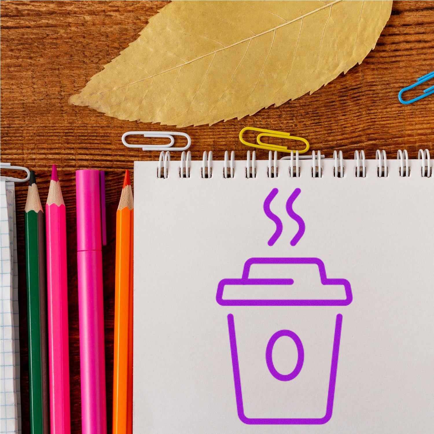 Self-Inking Java Joy Customer Loyalty Stamp with a purple coffee cup design on a notepad, surrounded by colorful pencils and paperclips on a wooden surface.