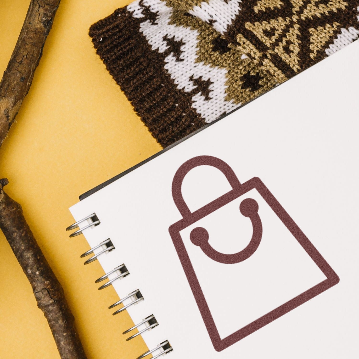 Self-Inking Shopper's Delight Membership Stamp with a shopping bag icon on a spiral notebook, set against a cozy background with a patterned knit and wooden sticks.