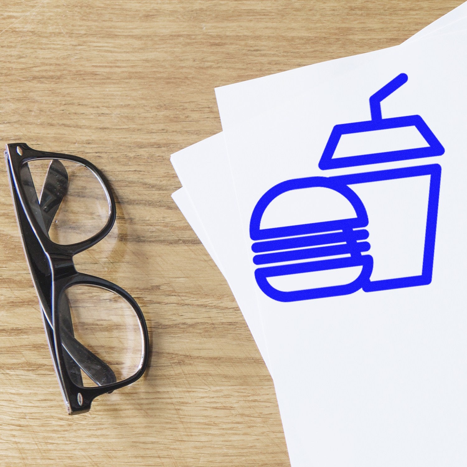 Image of the Self-Inking Burger Bliss Loyalty Punch Card Stamp, featuring a blue burger and drink icon on white paper, next to black glasses on a wooden surface.