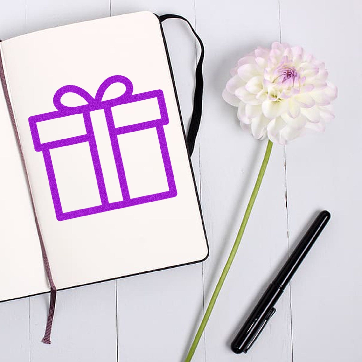 Self-Inking Gifts Galore Loyalty Points Stamp on a notebook, featuring a purple gift icon. Includes a black pen and a white flower on a light wooden surface. Perfect for rewards programs.