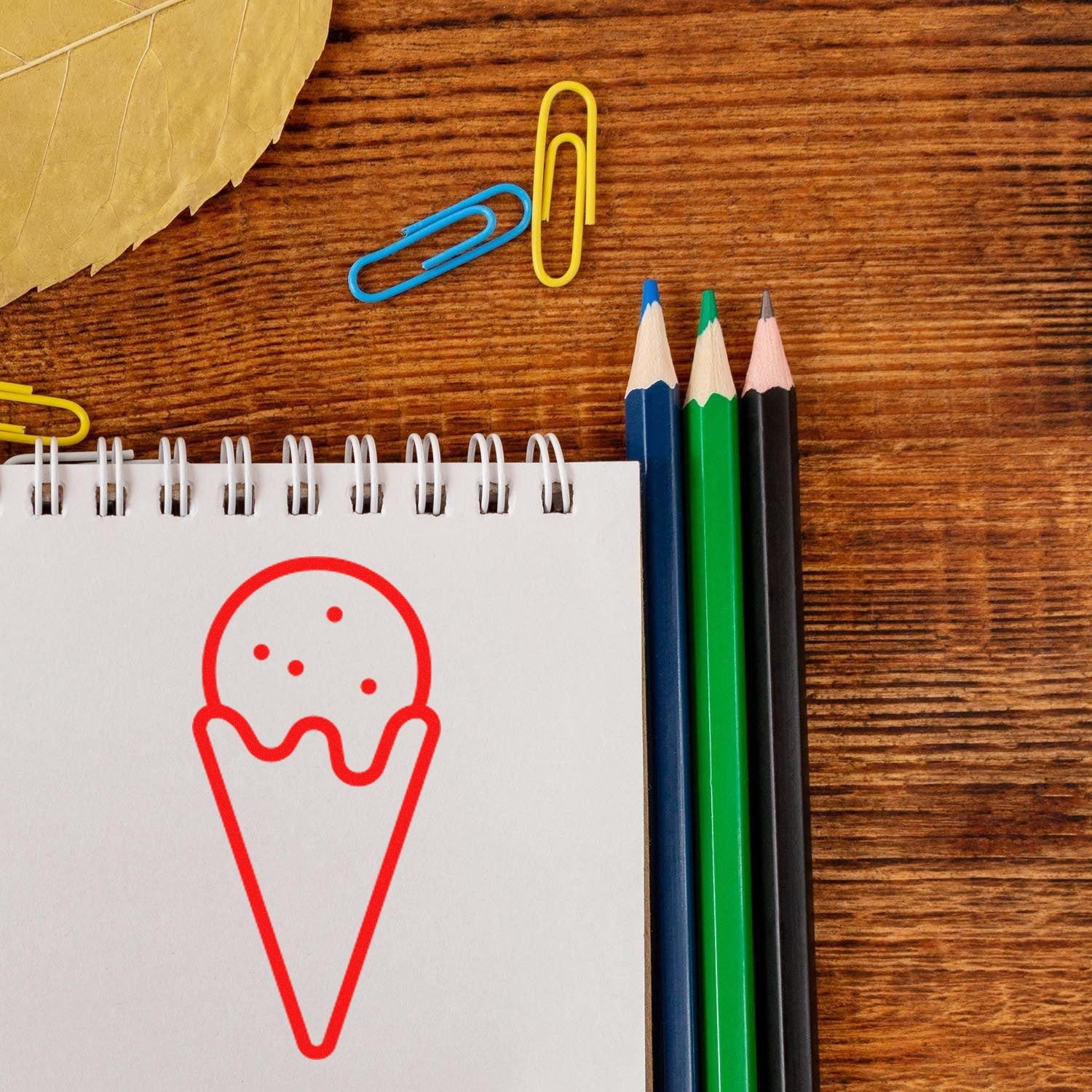 Wood Handle Sweet Rewards Purchase Rubber Stamp on a wooden surface, featuring a red ice cream cone design on paper, with colorful pencils and paperclips nearby.