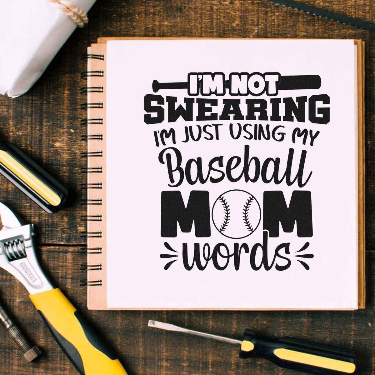 Custom stamp and seal design: black baseball mom words on a white notebook page, on rustic wood with yellow tools.