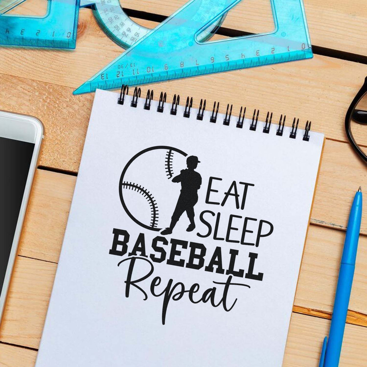 Black baseball stamp/seal design reading Eat Sleep Baseball Repeat on a white spiral notebook, with blue rulers on a wood desk.