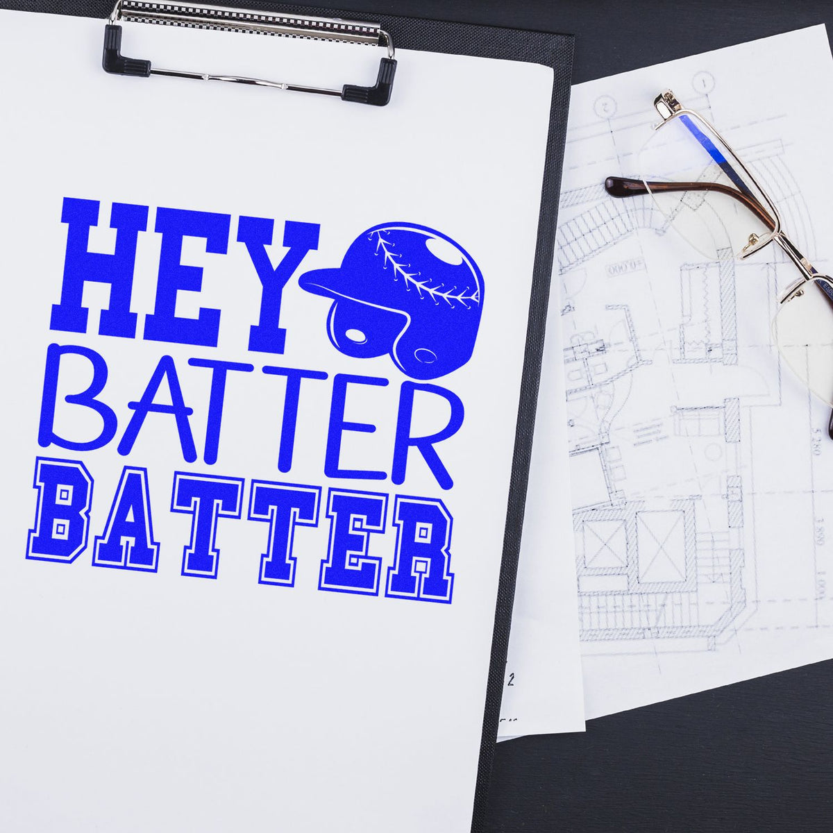 Blue custom stamp and seal design reading HEY BATTER BATTER with baseball helmet icon on clipboard, beside eyeglasses and blueprint sketches.