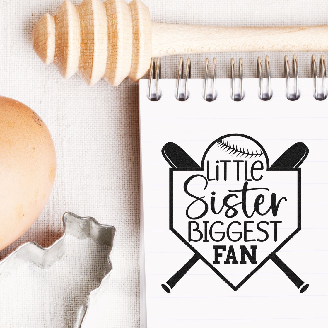 Black softball-themed stamp seal on a white spiral notebook reading Little Sister Biggest Fan, with a wooden handle and neutral craft items.