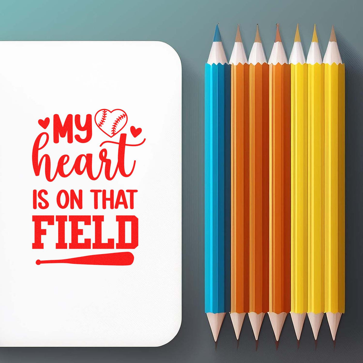 White custom stamp seal with red baseball-heart text, beside blue, orange, and yellow pencils on a gray background.