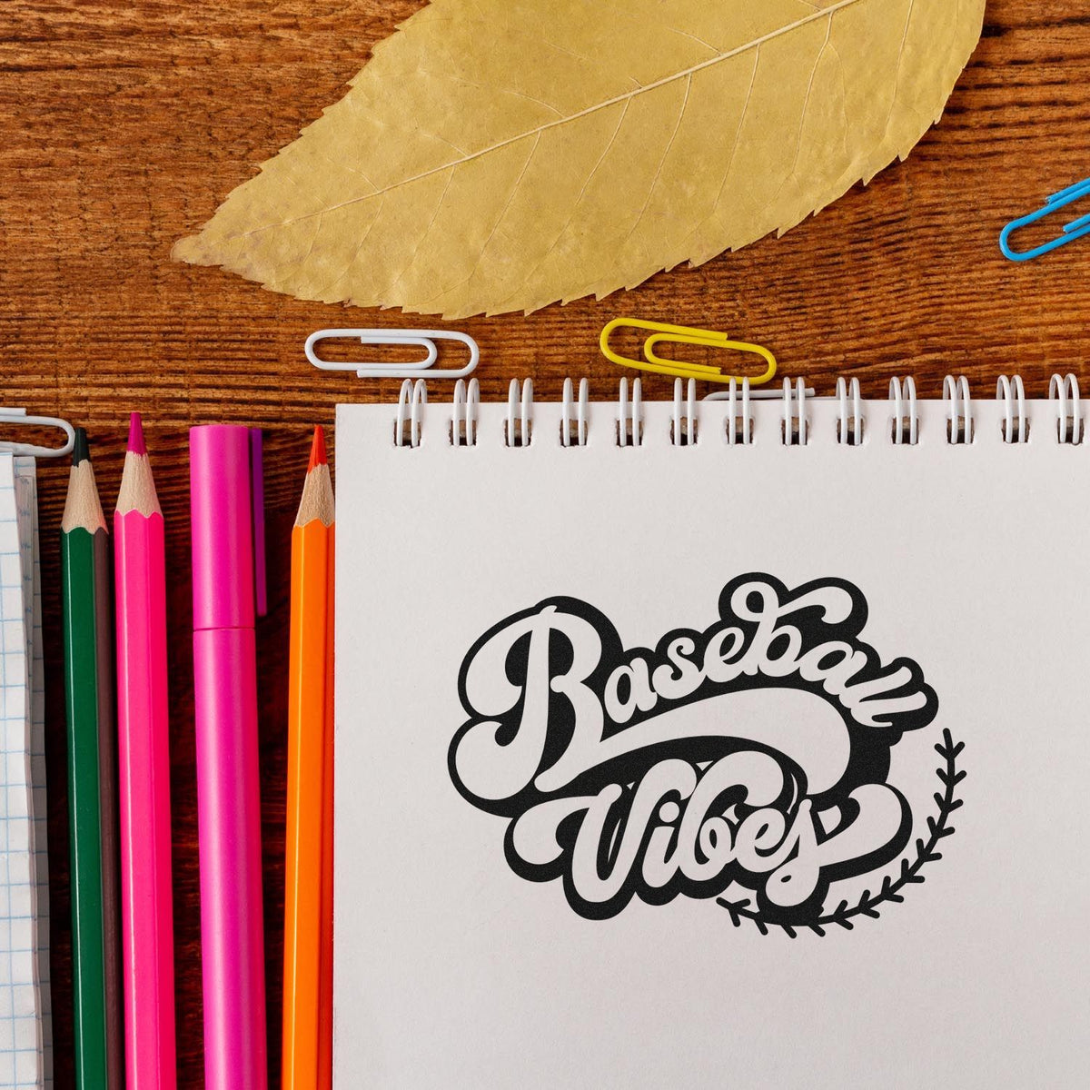 Black baseball-themed stamp seal on white spiral notebook, with colorful pencils, paper clips, and a beige leaf on wood.
