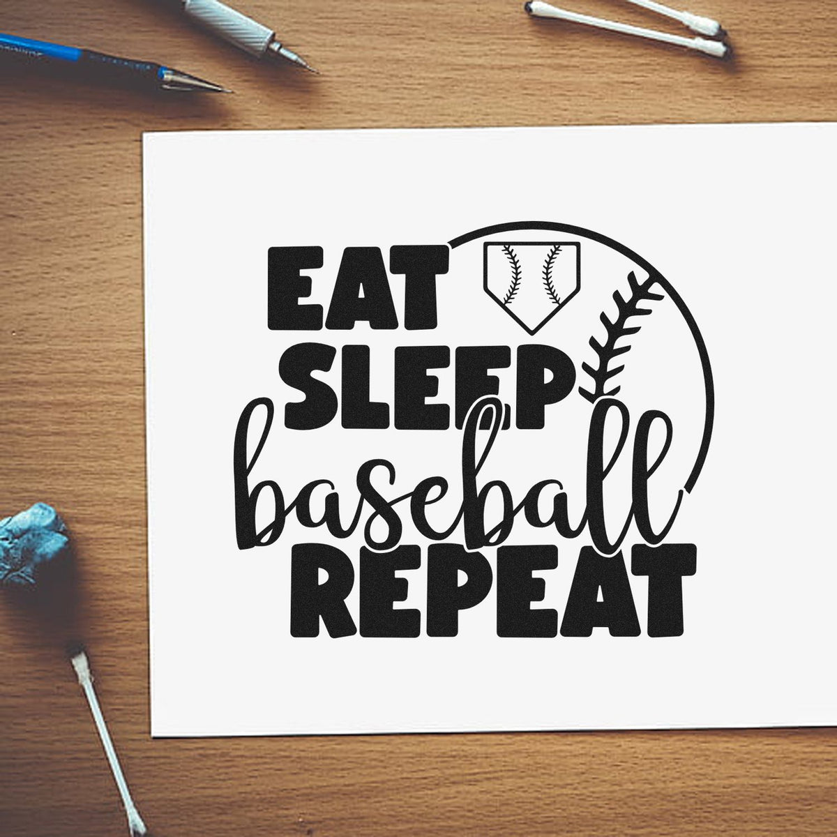 Black baseball rubber stamp and seal design reading EAT SLEEP baseball REPEAT on white paper over a wood desk.