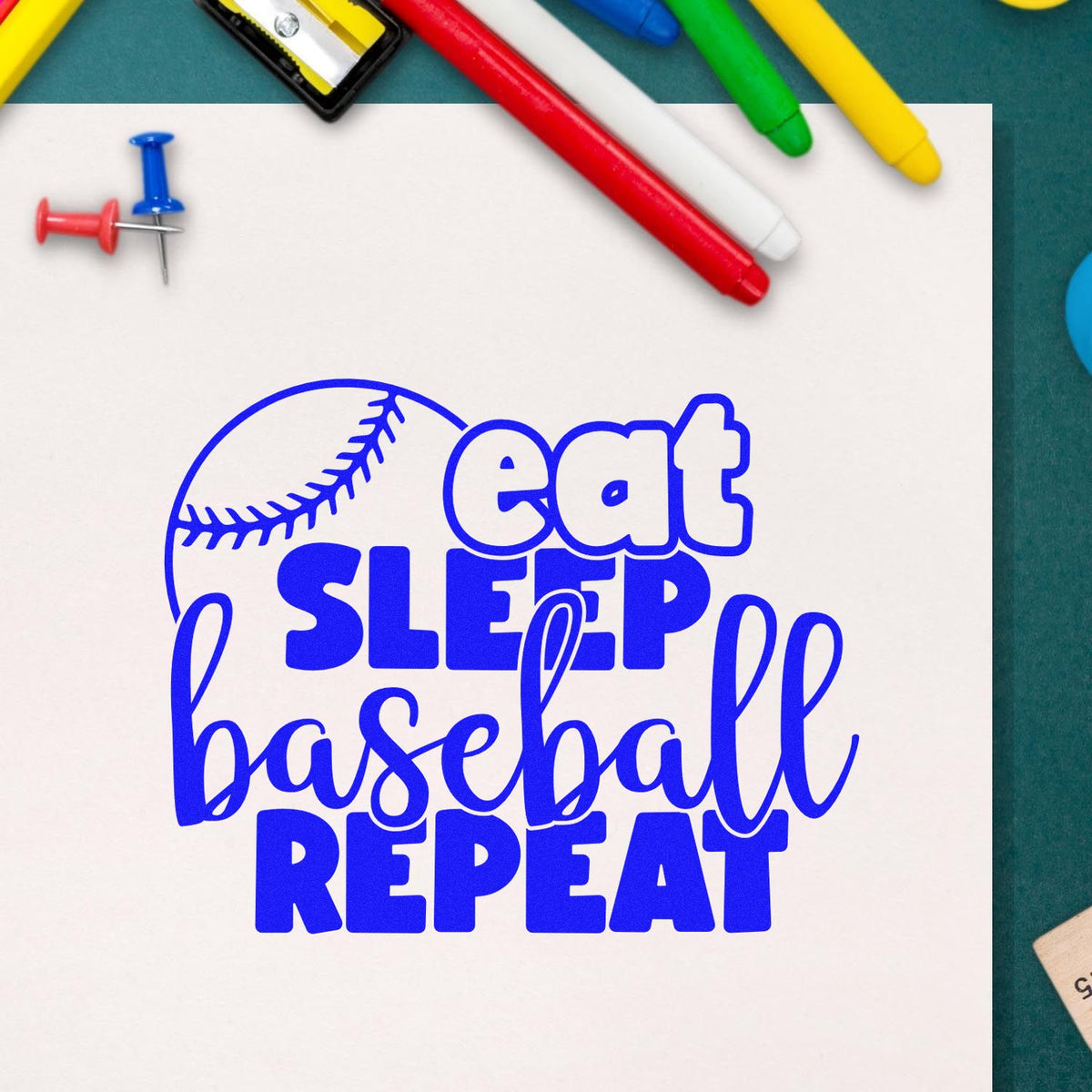 Blue baseball-themed stamp/seal on white paper reading eat sleep baseball repeat, surrounded by colorful markers and push pins.