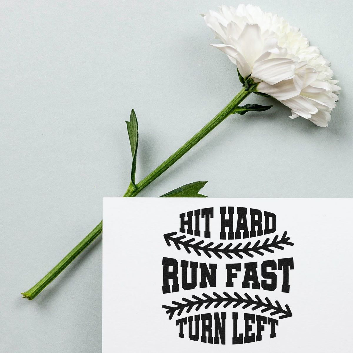 White flower beside a black custom rubber stamp and seal reading HIT HARD RUN FAST TURN LEFT on pale gray background.