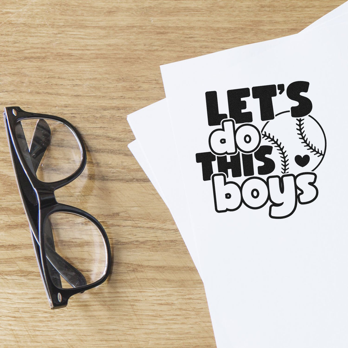 Black stamp design on white paper reading Lets do this boys with baseball graphics beside black eyeglasses on a wood table.