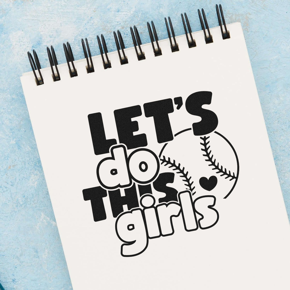 Black custom stamp and seal design reading Lets do this girls with baseball graphic on white spiral notebook, blue background.