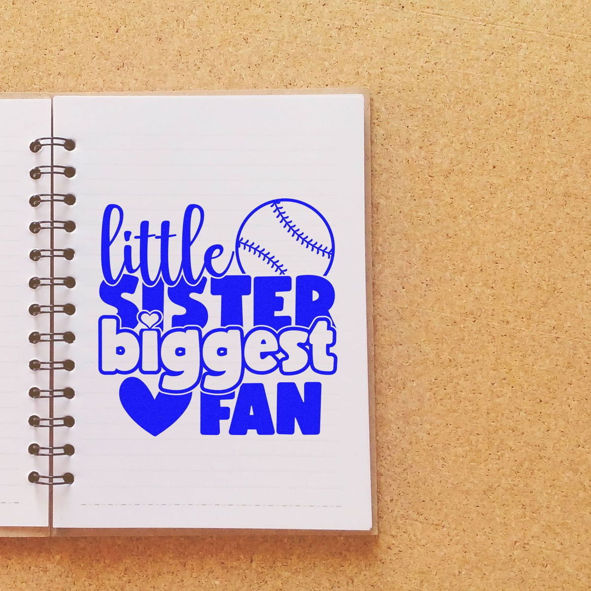 Blue custom stamp and seal reading little sister biggest fan with baseball and heart on a white spiral notebook.