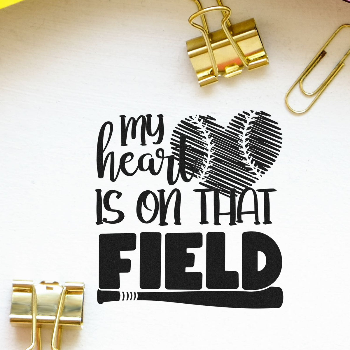 Black stamp design reading my heart is on that FIELD with a baseball heart graphic, on white paper with gold binder clips.
