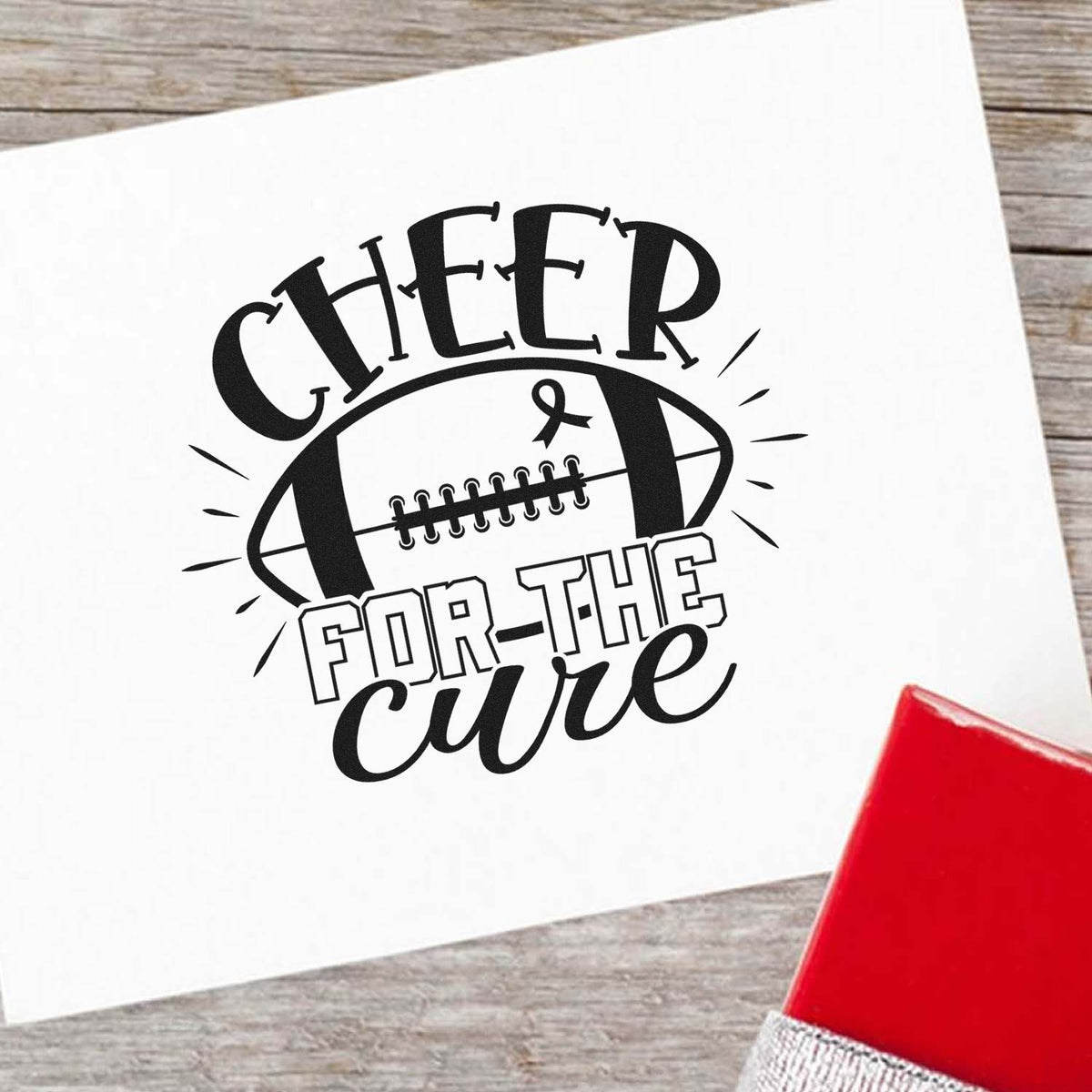 Black Cheer for the Cure football stamp seal on white paper, with a red stamp pad on rustic wood.