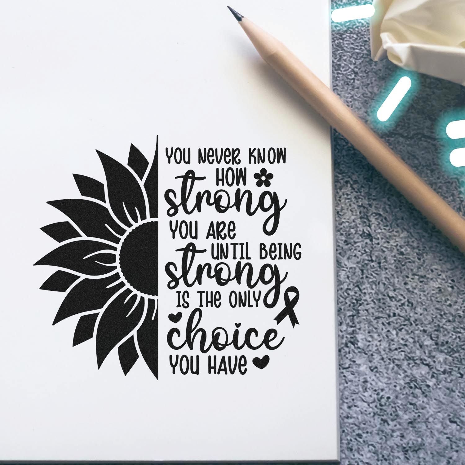 Black sunflower stamp and seal design with motivational quote on white paper, beside a tan pencil and ribbon accents.