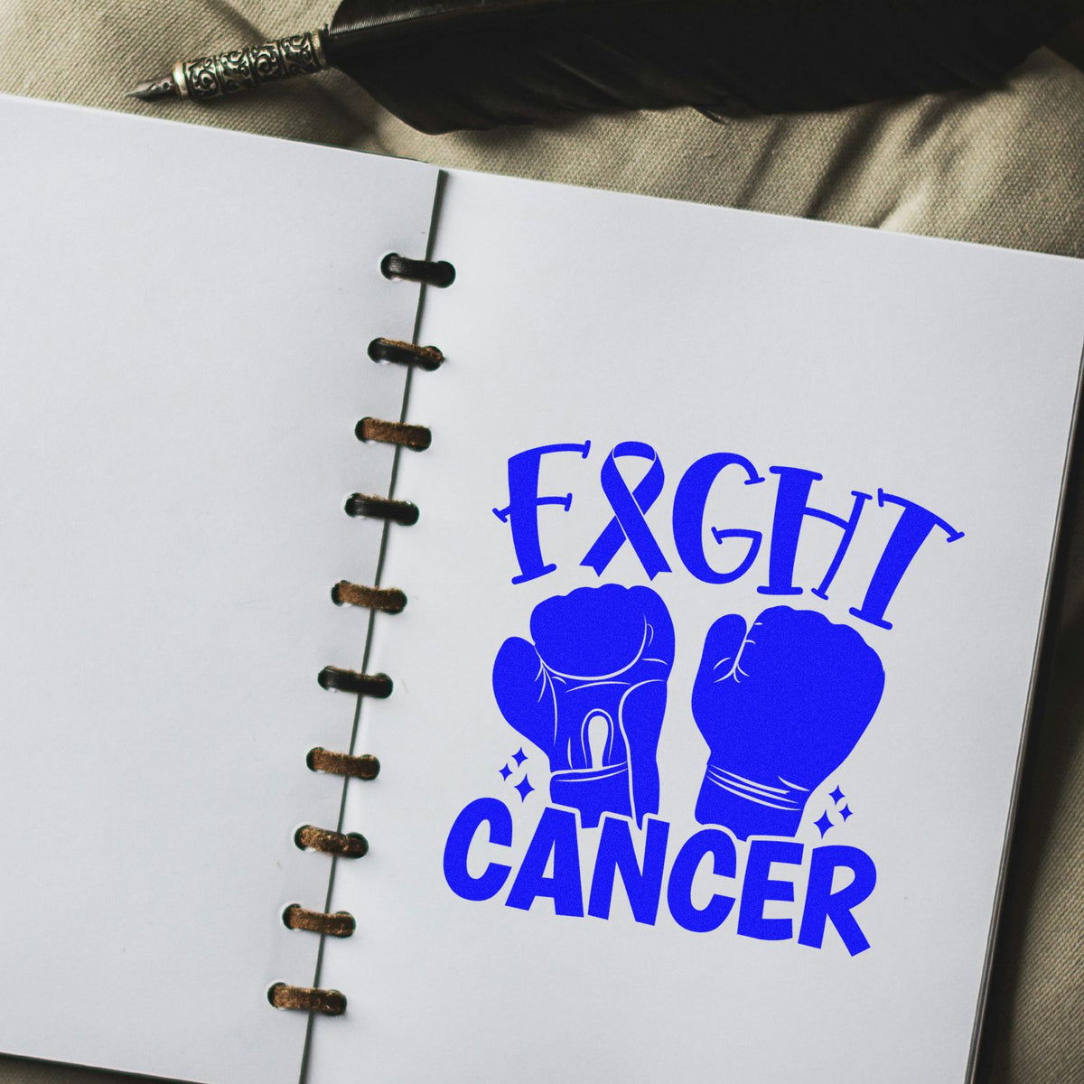 Blue Fight Cancer stamp and seal with boxing gloves on a white bound notebook, beside a black feather pen.