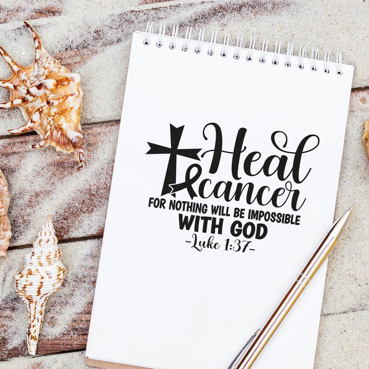 Black faith-based stamp and seal design on white spiral notebook, with seashells and gold pen on sandy beach.