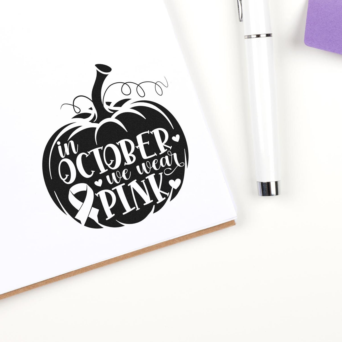 Black pumpkin stamp/seal with In October we wear pink on white card, beside white pen and purple envelope.