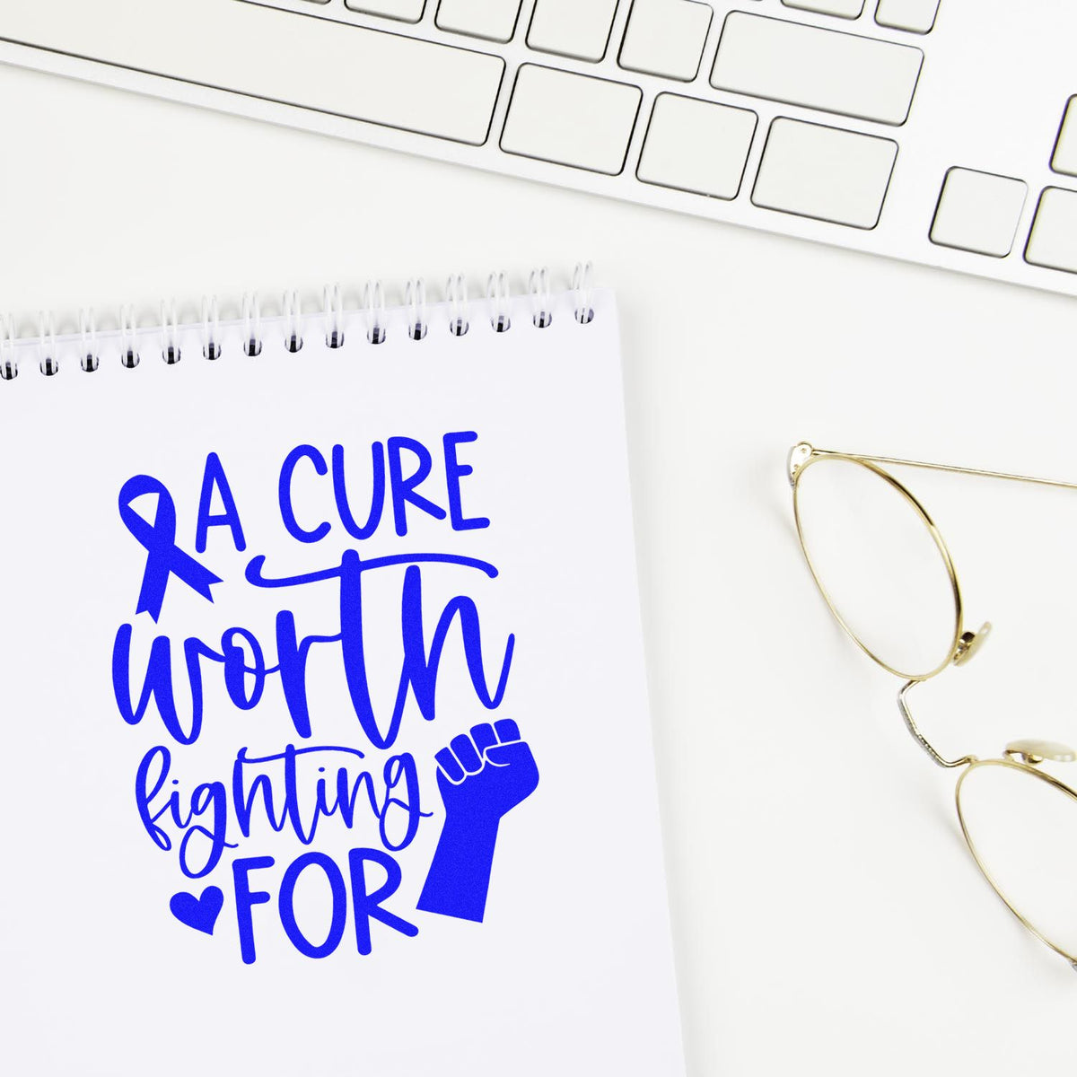 Blue breast cancer awareness stamp and seal design on white spiral notebook, with keyboard and gold glasses.