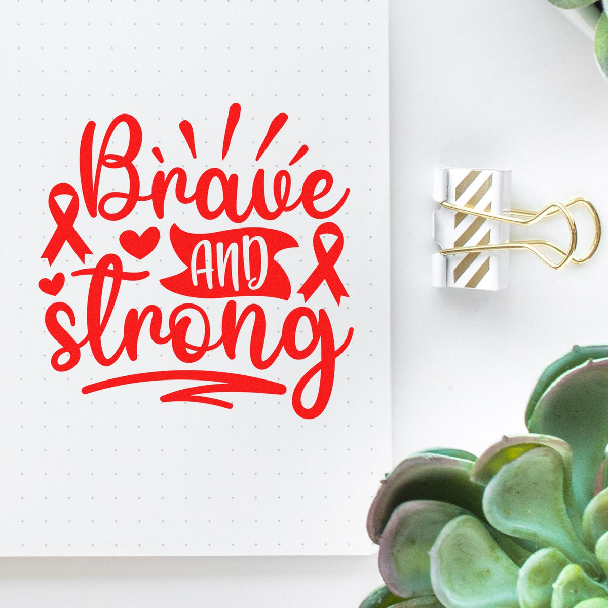 Red Brave and Strong stamp design on white dotted notebook page, clipped with a gold-striped binder clip beside a green succulent.