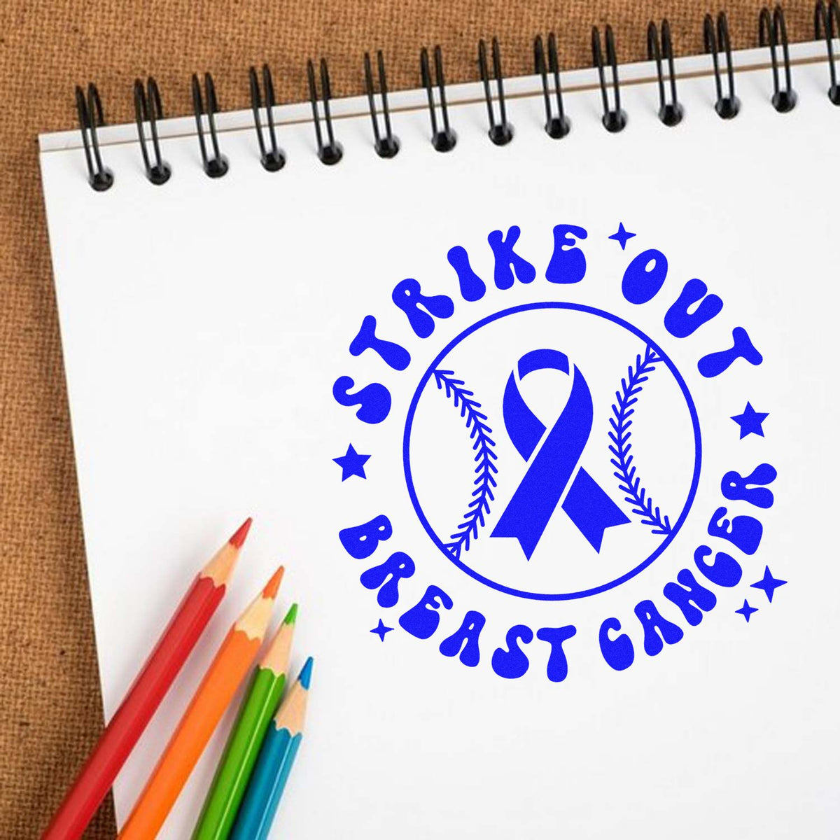 Blue breast cancer awareness stamp seal with ribbon and stars on a white spiral notebook, beside colorful pencils.